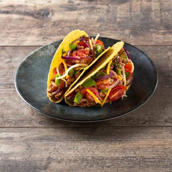 Two hard-shell tacos filled with ground meat, cheese, lettuce, tomatoes, and red onions on a dark plate.